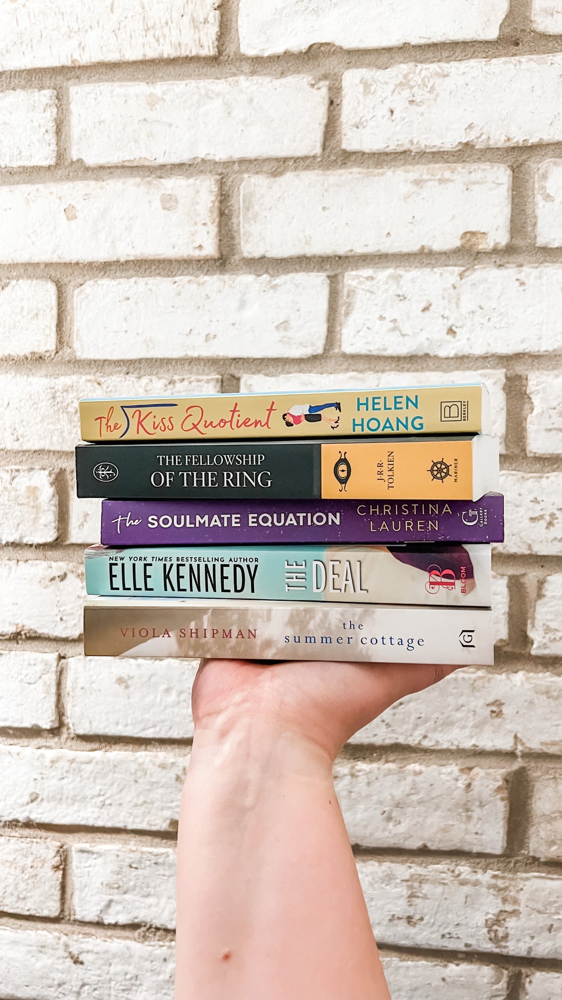 Photo of hand holding up a stack of books in front of a brick wall. The book titles are, from top to bottom, The Kiss Quotient, The Fellowship of the Ring, The Soulmate Equation, The Deal, and The Summer Cottage.